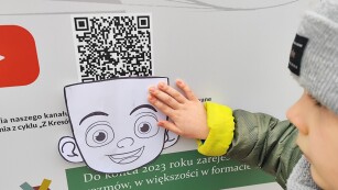 dziecko przykłada papierową twarz do znaku QR na plakacie